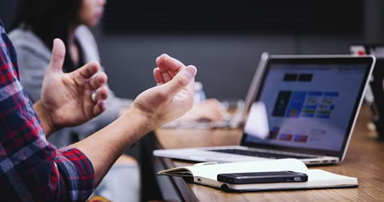 People in a meeting gesture with their hands, a laptop open on the table and a notebook nearby.